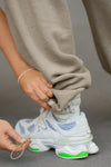 Close up detail side model from the knee down posing in the relaxed fit sand french terry Oversized Straight Leg Jogger with an elastic waistband, hidden drawstring, pocket details. and adjustable cinching at the ankle cuffs