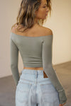 Back view of model posing in the fitted, slightly cropped sage modal Bare Shoulder Long Sleeve top with a straight, off-the-shoulder neckline