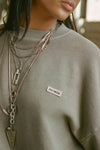 Close up detail front view of model posing in the oversized vetiver french terry Classic Crew Pullover sweatshirt with a crew neckline and joah brown logo patch sewn on the upper left chest