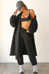 Full body front view of model posing in the knee-length loose fitting black french terry Oversized Cardigan with a rolled neckline and extra long sleeves