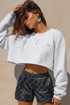 Front view of model posing in the loose fit cropped pearl grey french terry Cut Off Sweatshirt with oversized long sleeves, a raw cut hem and a white patch with a black joah brown logo on it sewn on the upper left chest