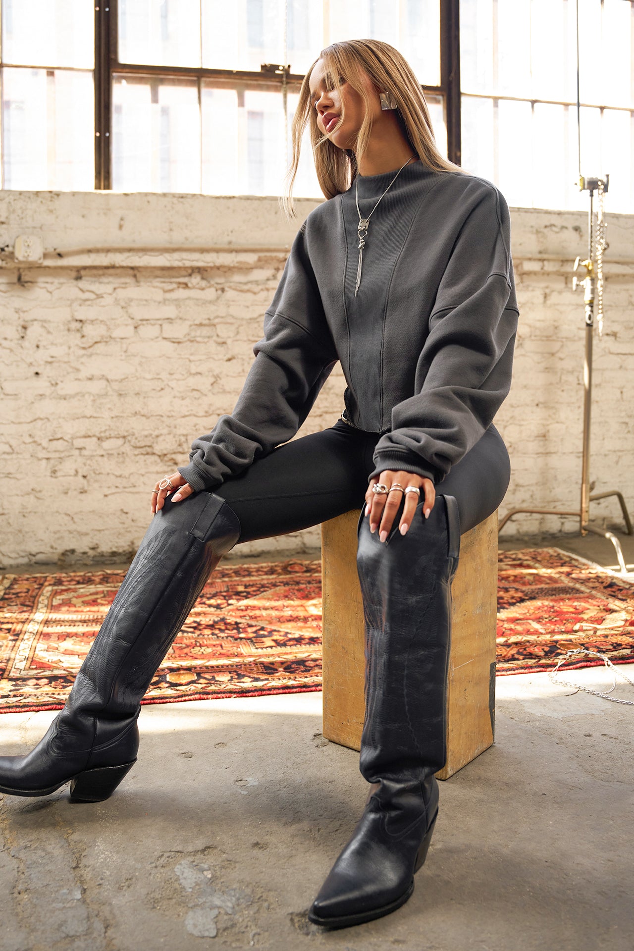 Full body view of model sitting on a box and posing in the charcoal luxe french terry Dolman Pullover sweatshirt with oversized sleeves, a mock neckline, curved hem and ribbed panels at the front and back