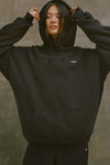 Front view of model posing in the oversized comfortable black french terry Oversized Pullover Hoodie sweatshirt with a Joah Brown logo patch at the front left chest, kangaroo pocket, drawstrings and thumbholes