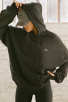 Front view of model posing in the oversized comfortable black french terry Oversized Pullover Hoodie sweatshirt with a Joah Brown logo patch at the front left chest, kangaroo pocket, drawstrings and thumbholes