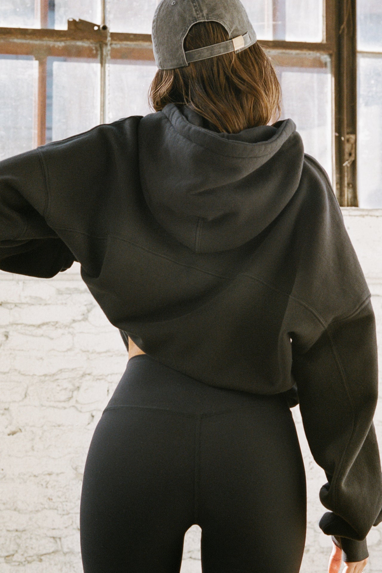 Back view of model posing in the oversized comfortable black french terry Oversized Pullover Hoodie sweatshirt with paneling on the back and arms and thumbholes