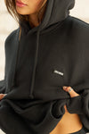 Close up detail front view of model posing in the oversized comfortable black french terry Oversized Pullover Hoodie sweatshirt with a Joah Brown logo patch at the front left chest, kangaroo pocket, drawstrings and thumbholes