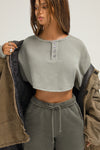 Front view of model posing in the relaxed fit vetiver french terry Henley Crop Sweatshirt with distressed cuffs and placket, dropped shoulders and custom JOAH BROWN buttons