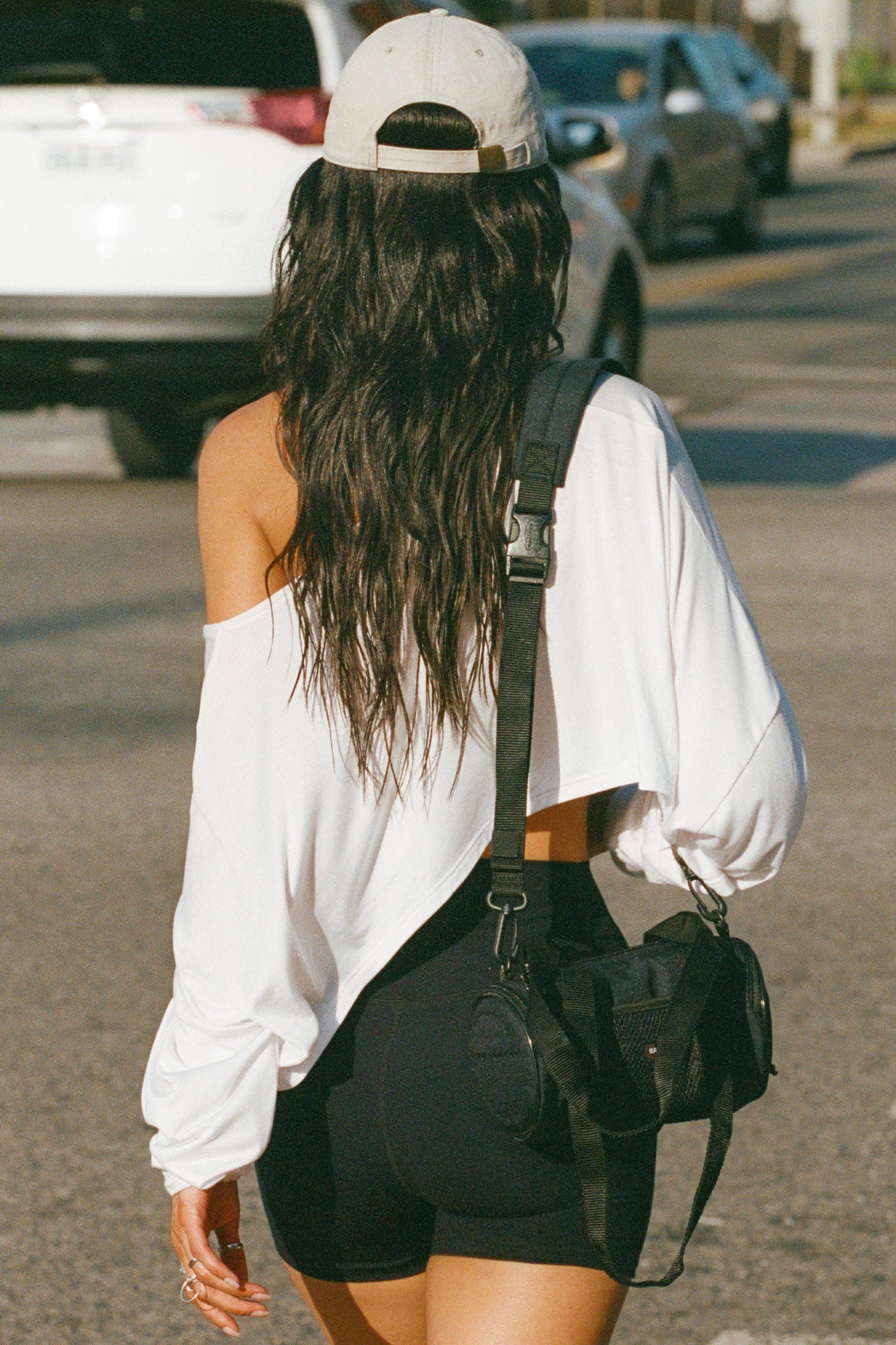 Back view of model posing in the street wearing the cropped, loose fitting white modal Relaxed Crop Long Sleeve top with a wide scoop neckline that can be worn off the shoulder