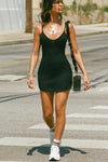 Front view of model walking in the form fitting stretchy black rib mini Slip Dress with thin straps, a u neckline and a tiny side slit at the hem