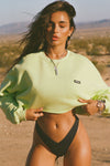 Front view of model posing in the loose fit cropped melon french terry Cut Off Sweatshirt with oversized long sleeves, a raw cut hem and a white patch with a black joah brown logo on it sewn on the upper left chest