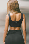 Back view of model posing in the fitted sueded onyx Scoop Neck Smoothing Bra top with a scoop back and Joah Brown logo patch that can be worn in the front or the back