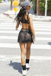 Full body back view of model walking in the mini black mesh skirt with an adjustable waist tie