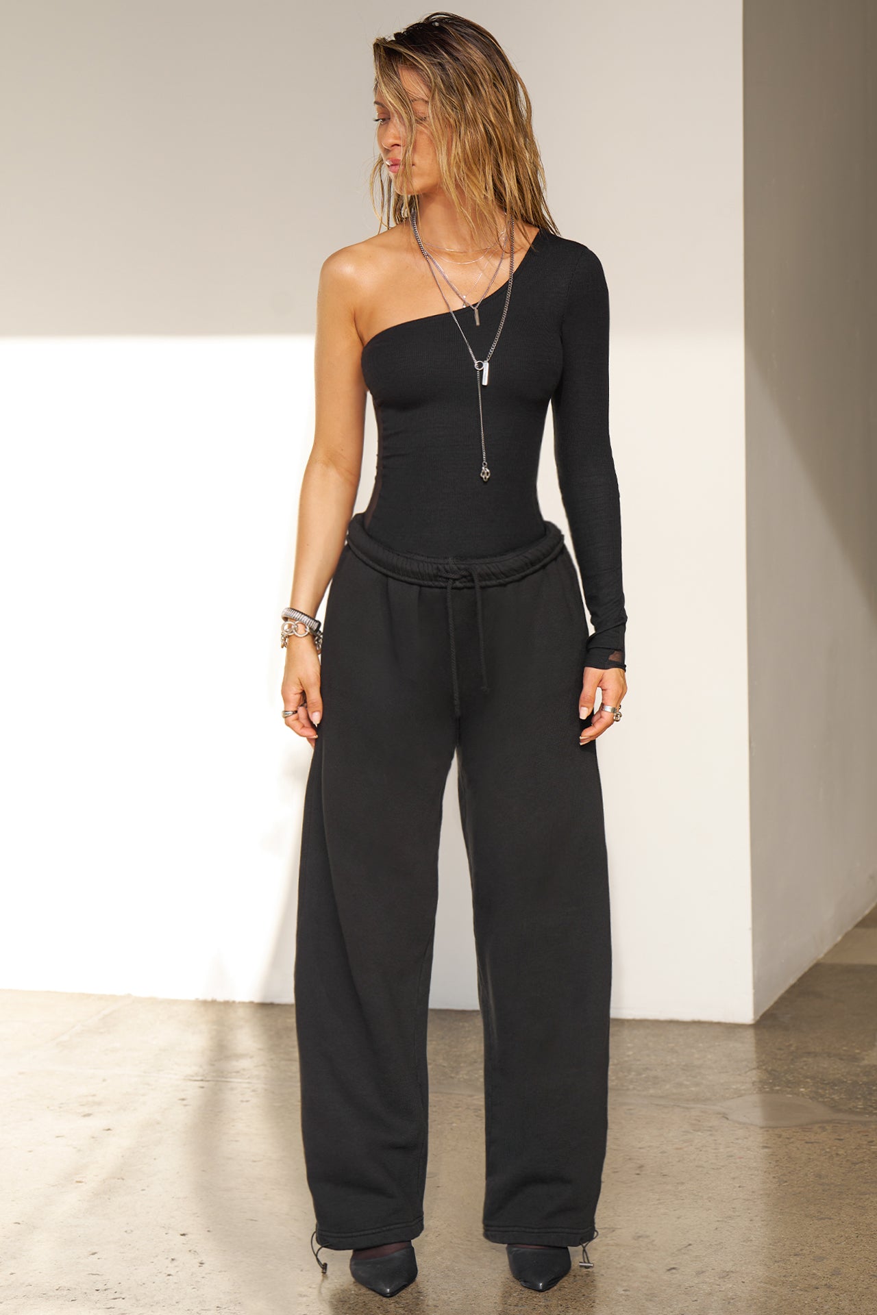 Full body front view of model posing in he fitted black flexrib One Shoulder Long Sleeve top with an asymmetrical neckline, flexrib front and mesh back.