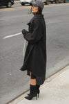 Full body side view of model posing in the knee-length loose fitting black french terry Oversized Cardigan with a rolled neckline and extra long sleeves