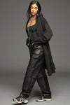 Full body side view of model posing in the knee-length loose fitting black french terry Oversized Cardigan with a rolled neckline and extra long sleeves