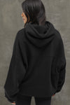 Back view of model posing in the oversized comfortable black french terry Oversized Pullover Hoodie sweatshirt with paneling on the back and arms and thumbholes