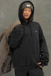 Front view of model posing in the oversized comfortable black french terry Oversized Pullover Hoodie sweatshirt with a Joah Brown logo patch at the front left chest, kangaroo pocket, drawstrings and thumbholes