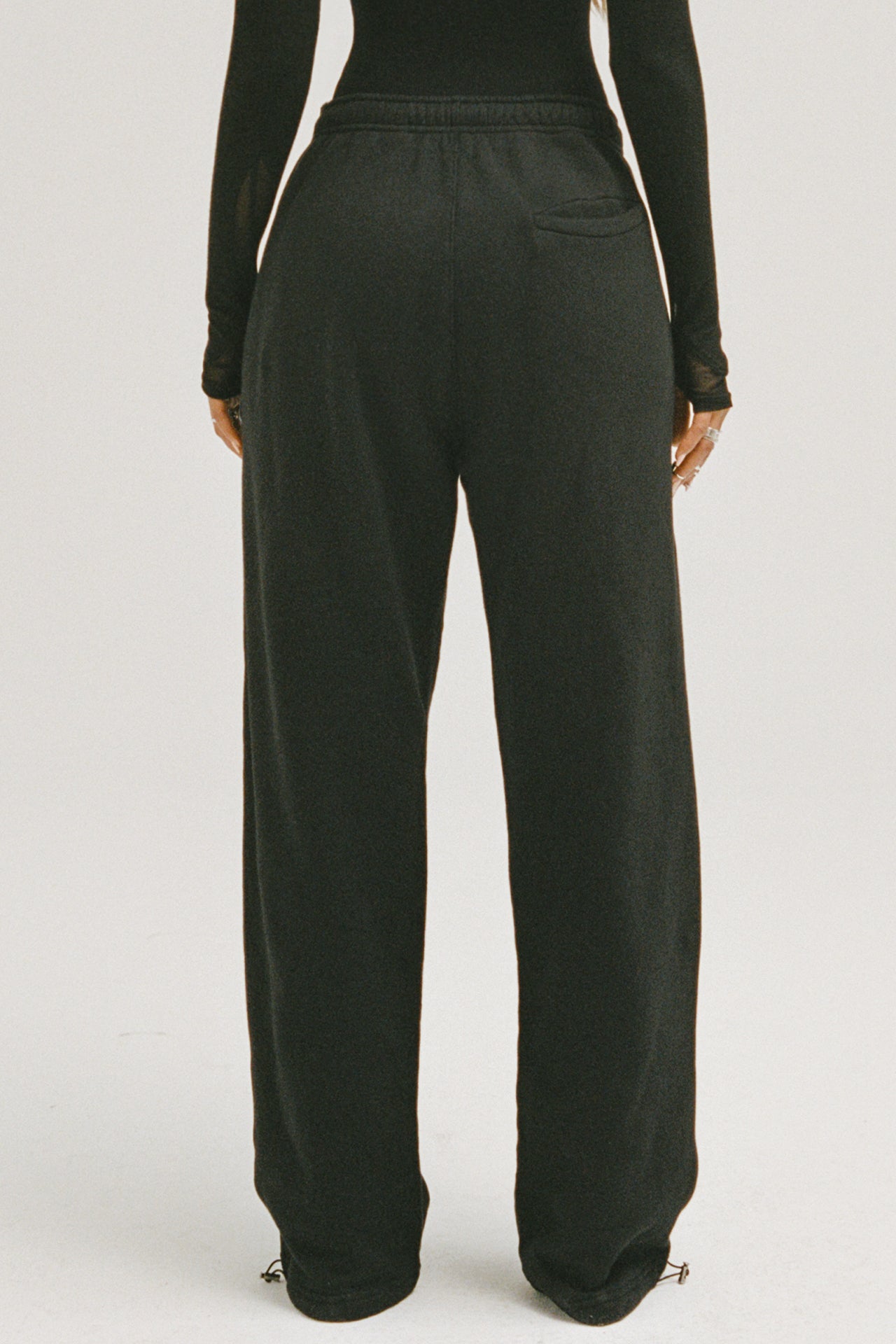 Back view of model from the waist down posing in the relaxed fit black french terry Oversized Straight Leg Jogger with an elastic waistband, hidden drawstring, pocket details. and adjustable cinching at the ankle cuffs