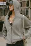 Front view of model posing outside in the street in the oversized comfortable vetiver french terry Oversized Zip Hoodie with a full length front zip closure, side pockets and thumbholes in the fitted cuffs