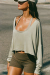 Side view of model posing in the street wearing the cropped, loose fitting sage modal Relaxed Crop Long Sleeve top with a wide scoop neckline that can be worn off the shoulder