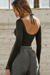 Back view of model posing in the fitted black flexrib Scoop Corset Long Sleeve with a low scoop back, adjustable ties at the neck, and thumbholes in the mesh sleeves