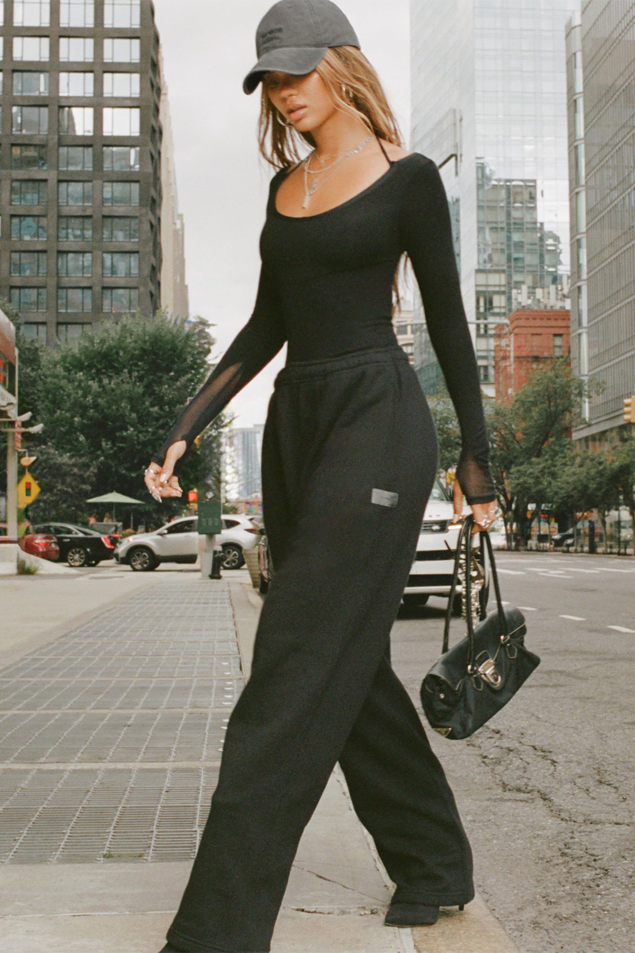 Full body side view of model posing in the street posing in the fitted black flexrib Scoop Corset Long Sleeve with a scoop neckline, adjustable ties at the neck, and thumbholes in the mesh sleeves